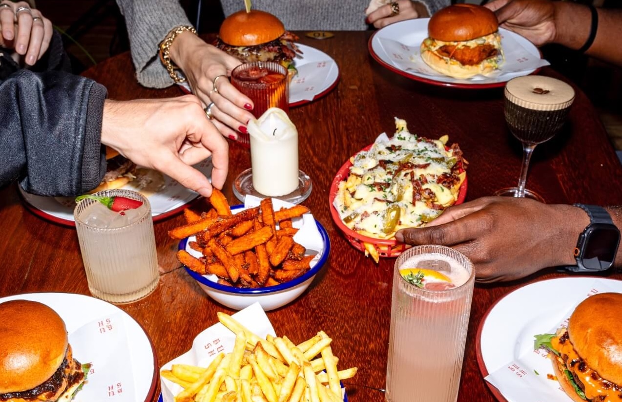 People tucking into a table full of burgers, chips and cocktails
